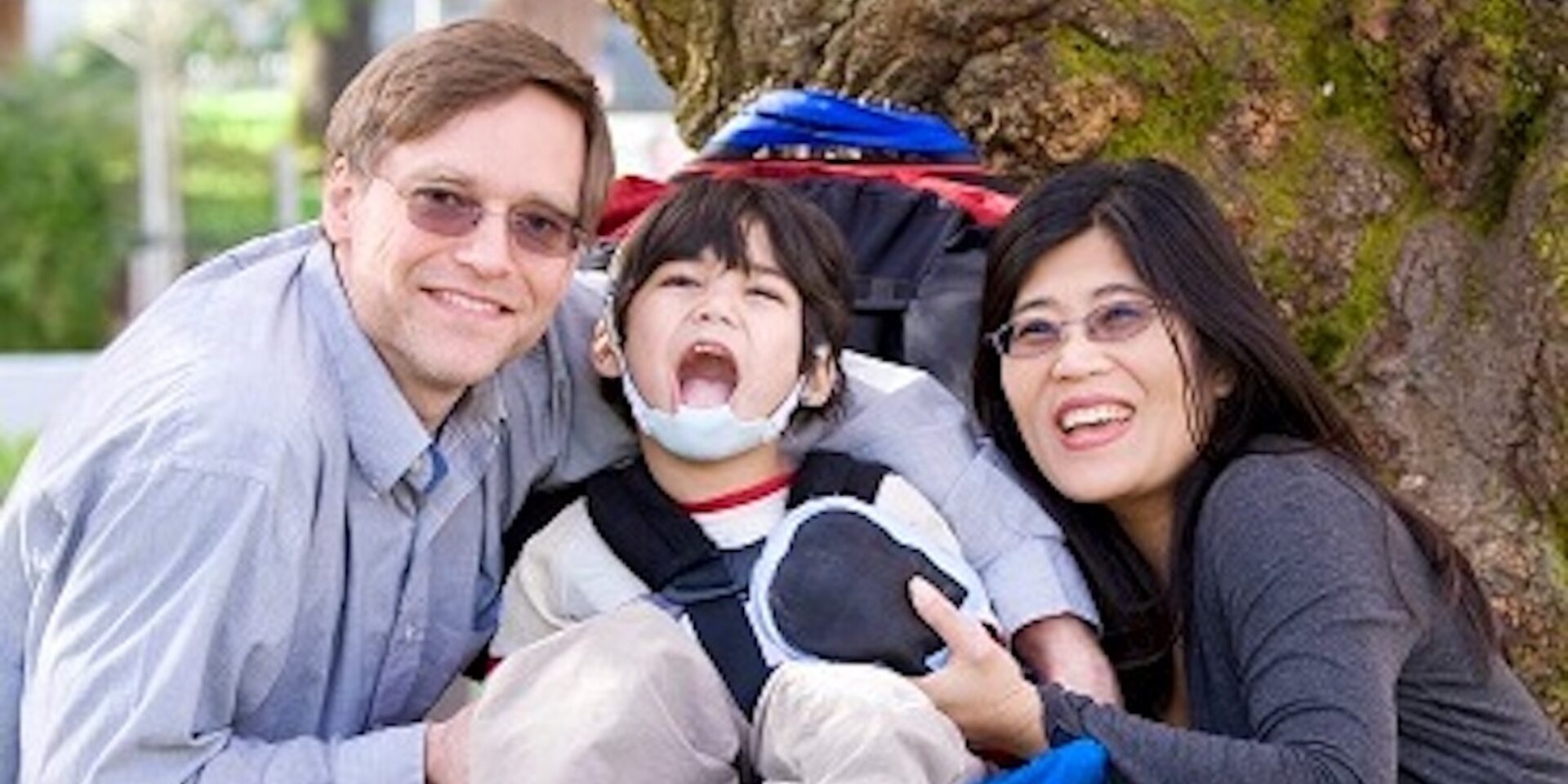 A photo of a Mum and Dad with their son who is in a wheelchair, out in the fresh air together next to a tree.