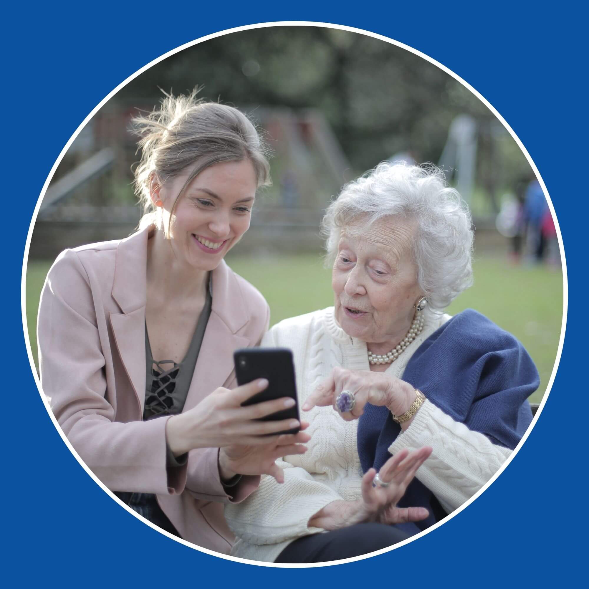 Photo showing a younger lady sitting with an elderly lady in a park, smiling looking at a phone. Image from Pexels (Photographer - Andrea Piacquadi)