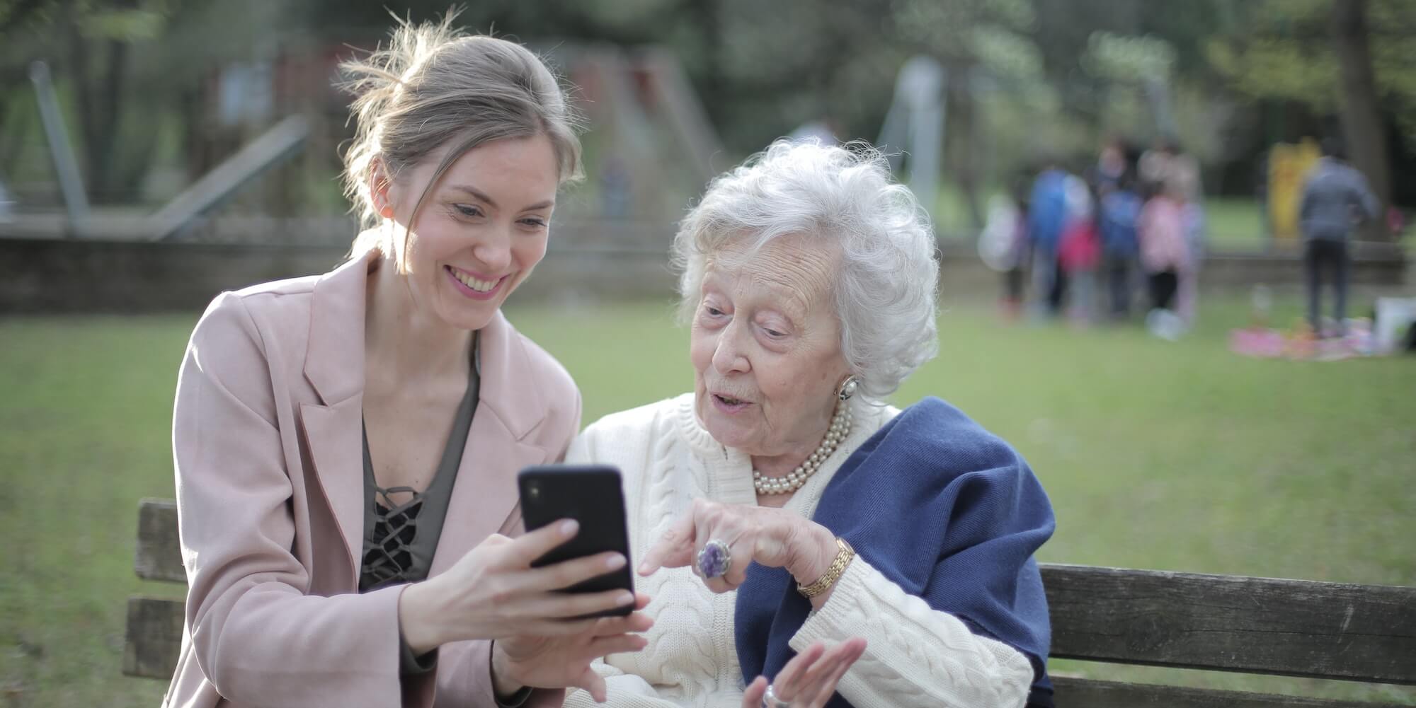 Photo showing a younger lady sitting with an elderly lady in a park, smiling looking at a phone. Image from Pexels (Photographer - Andrea Piacquadi)