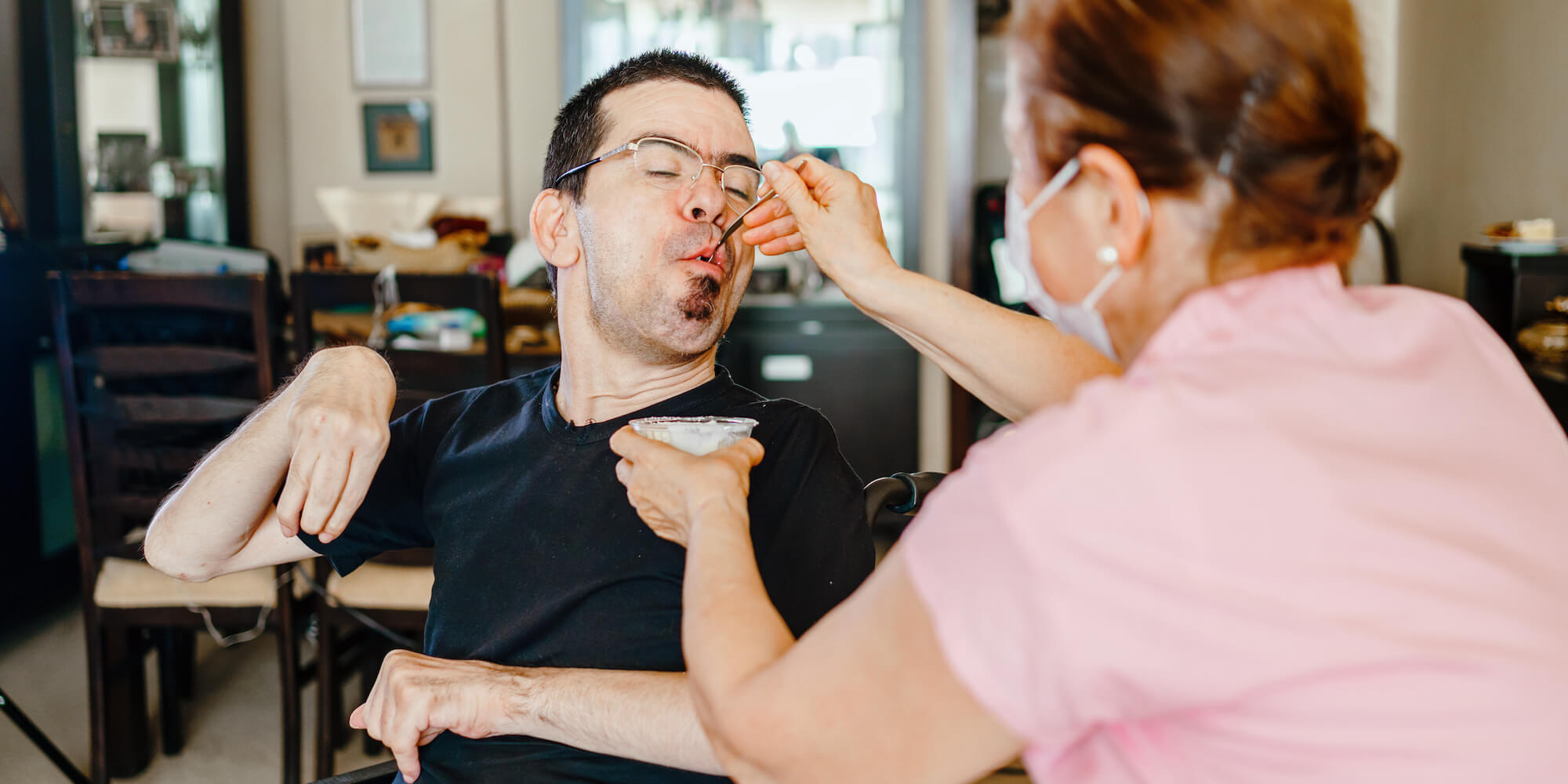 A Personal Assistant helping to feed a gentleman with a disability in a wheelchair (Image from iStock-1255038289)