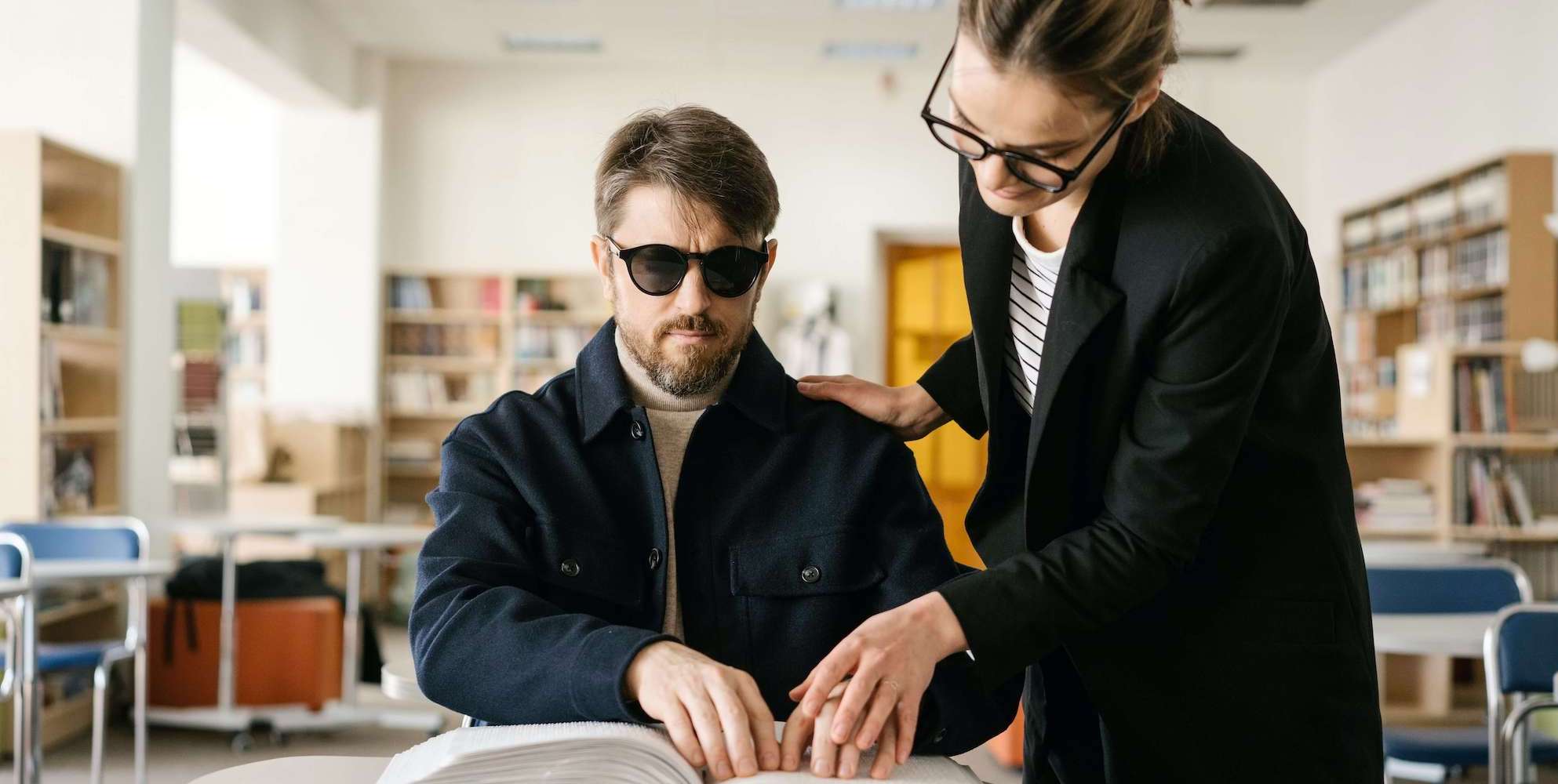 A lady helping a blind gentleman read a braille book in a library. Photo from Pexels (Photographer is Mikhail Nilov)