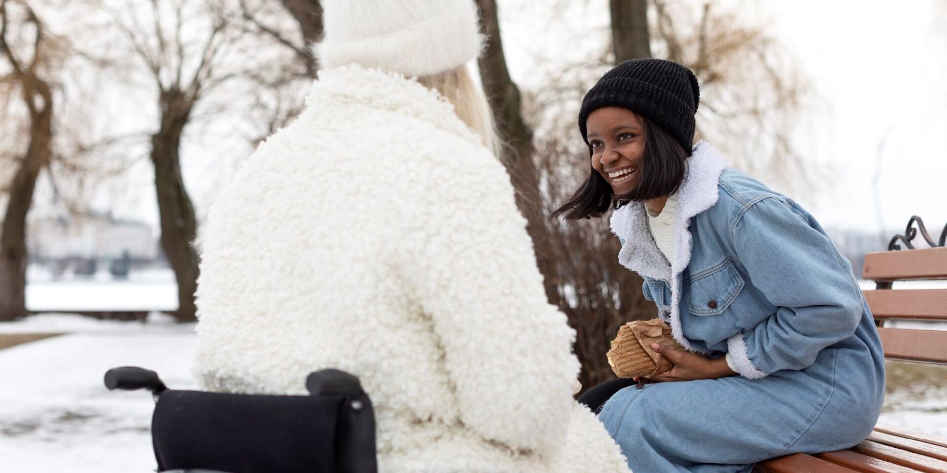 A photo showing a lady in a wheelchair and a smiling girl sitting on a park bench, outside enjoying lunch in the snow.